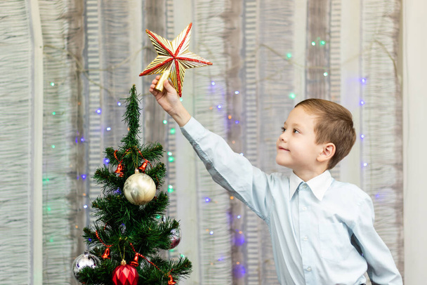 Yedi yaşında gömlekli bir çocuk Noel ağacını Noel arifesinde evde çelenklerle süslüyor. Seçici odaklanma. Portre - Fotoğraf, Görsel