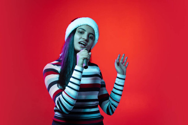 Portrait of young cute girl, student in warm clothes singing, using microphone isolated on red color studio background - Valokuva, kuva