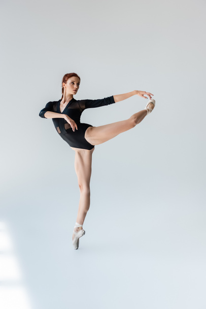 full length of flexible ballerina in black bodysuit stretching on grey - Fotografie, Obrázek
