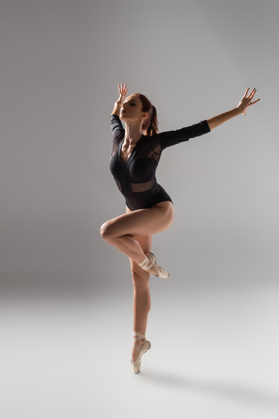 full length of flexible ballerina in black bodysuit dancing on dark grey - Fotografie, Obrázek