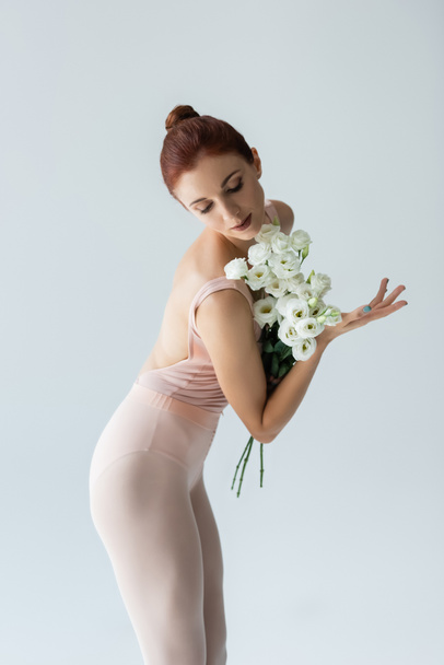 redhead ballerina with closed eyes holding bouquet of eustoma flowers isolated on grey  - Photo, image