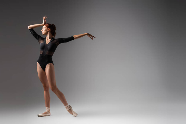 volle Länge der anmutigen Ballerina im schwarzen Body stehend mit ausgestreckter Hand auf dunkelgrau - Foto, Bild