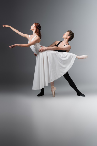 full length of graceful ballerina in white dress dancing with young partner on dark grey - 写真・画像