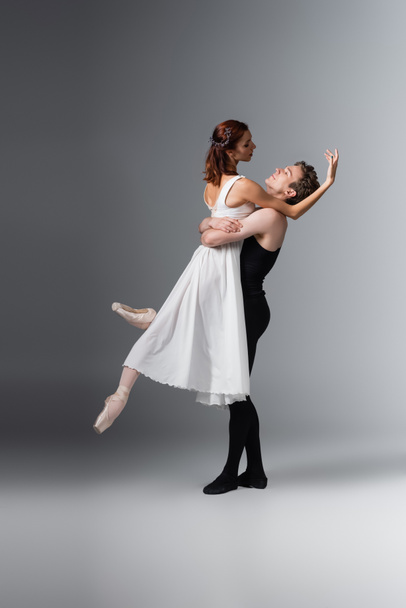 full length of dancer lifting graceful ballerina in white dress on dark grey - Foto, afbeelding