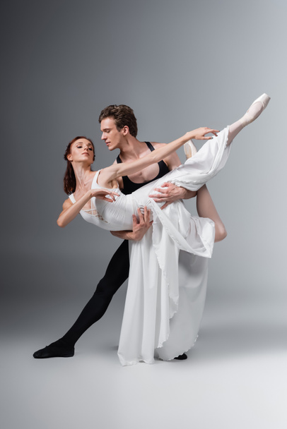 full length of man supporting graceful ballerina in white dress while dancing on dark grey - Foto, imagen