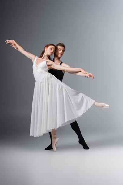 full length of graceful ballerina in white dress dancing with young partner on dark grey - Foto, immagini