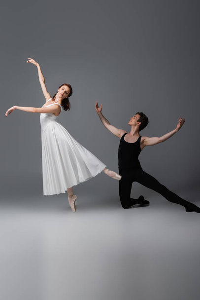 полная длина изящной балерины в белом платье танцует рядом с партнером стоя на коленях на темно-серой - Фото, изображение