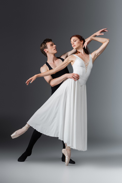 volle Länge der jungen anmutigen Ballerina im weißen Kleid, die mit Partner auf dunkelgrau tanzt - Foto, Bild