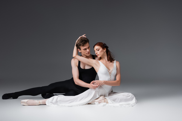 full length of flexible dancer and graceful ballerina in white dress sitting on dark grey - Zdjęcie, obraz