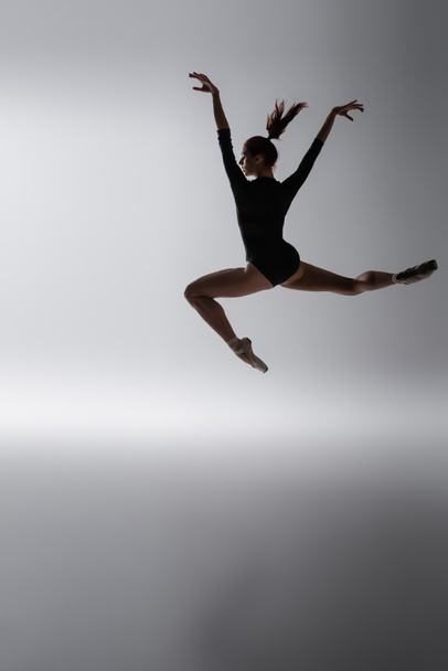 redhead ballerina in bodysuit jumping with outstretched hands on dark gray - Foto, Imagem