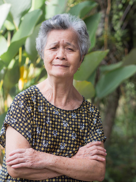 Портрет пожилой азиатки с короткими белыми волосами, в очках, улыбающейся с скрещенными руками, и смотрящей в камеру, стоя в саду. Концепция пожилых людей и здравоохранения. - Фото, изображение