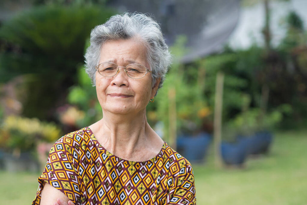 Пожилая азиатка в очках, улыбающаяся и смотрящая в камеру, стоя в саду. Пространство для текста. Концепция пожилых людей и здравоохранения. - Фото, изображение
