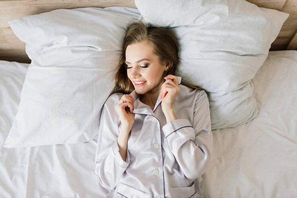 Фото молодой веселой женщины в пижаме, печатающей на ноутбуке и улыбающейся, сидя на кровати в светлом помещении - Фото, изображение