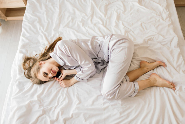 Foto von jungen fröhlichen Frau im Pyjama tippt auf Laptop und lächelt, während sie auf dem Bett im hellen Zimmer sitzt - Foto, Bild