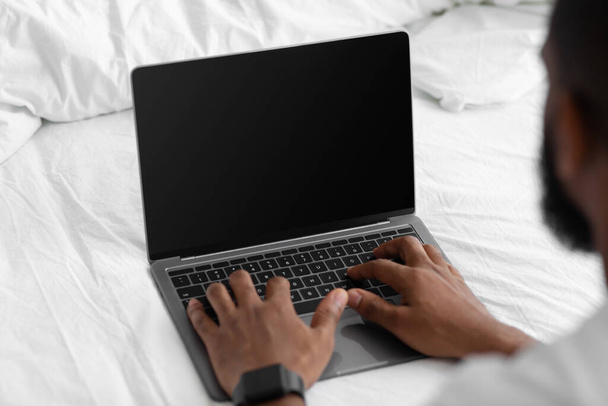 Młody Afroamerykanin patrzy na laptopa z pustym ekranem, pracuje zdalnie na białym łóżku w sypialni - Zdjęcie, obraz