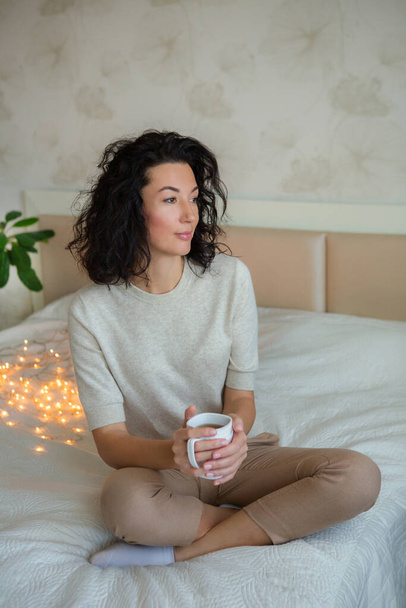 Young woman holding a cup of tea in bed. Christmas lights on, cozy home atmosphere. - Φωτογραφία, εικόνα