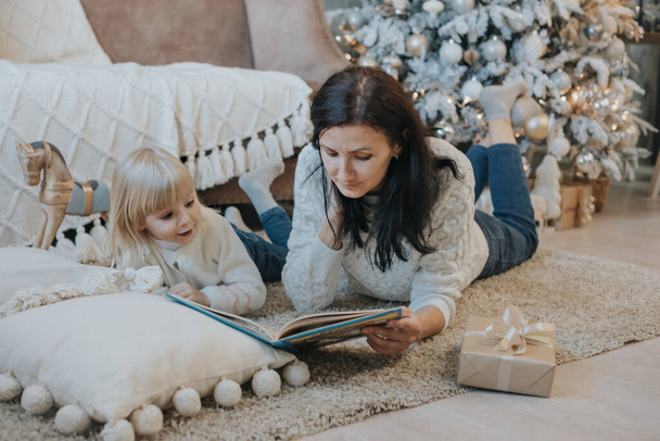 Mom and daughter are reading a book lying on the carpet near the Christmas tree - Foto, immagini