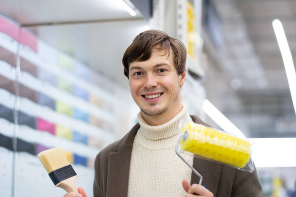 attraktiver europäischer Mann hält Farbwalze und Pinsel in der Hand in einem Baumarkt - Foto, Bild