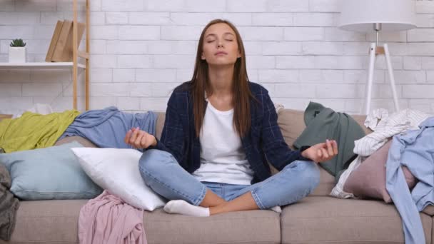 Prácticas domésticas y sin estrés. Joven tranquila mujer pacífica meditando entre la ropa dispersa, sentado en el sofá - Imágenes, Vídeo