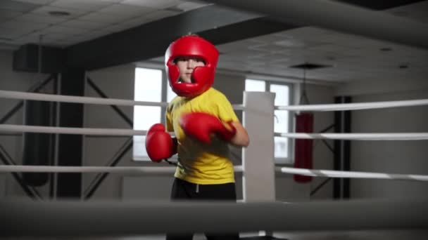 Mały chłopiec w rękawiczkach bokserskich i miękkim hełmie na głowie i pokazuje swoje ruchy - Materiał filmowy, wideo