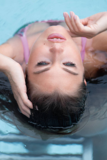Από πάνω σαγηνευτική γυναίκα αγγίζει βρεγμένα μαλλιά και δίχτυ, ενώ στηρίζεται σε καθαρό νερό της πισίνας στο θέρετρο - Φωτογραφία, εικόνα