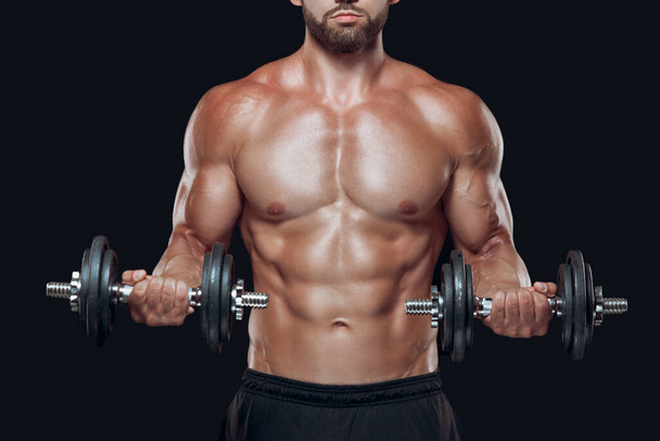 Close up of muscular body and strong hands lifting heavy dumbbells isolated over black background - Foto, afbeelding