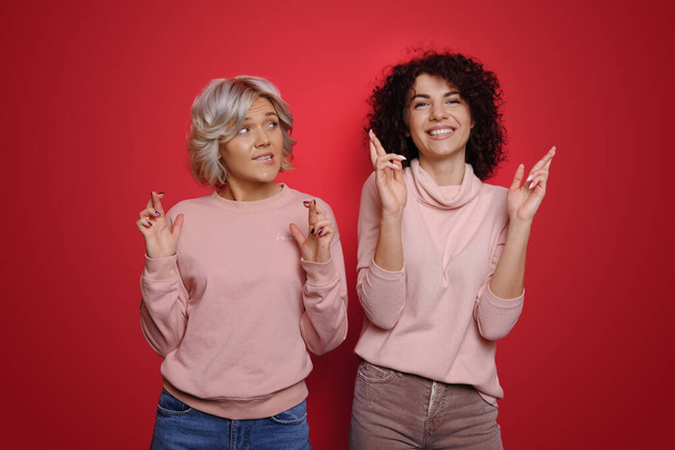 Photo of two women holding crossed fingers wishing luck success isolated over red color background. Curly hair - Photo, Image