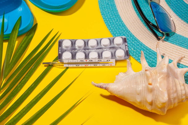 Top view on white pills in blister pack, thermometer, blue hat, blue flip-flops, sunglasses, seashell, palm leaf on yellow background. Concept of beach holiday, sea tour, warm sunny summer - Foto, afbeelding