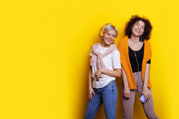 Two women friends in casual t-shirts listening music with headphones dancing isolated on yellow wall background with copyspace. Blank space - Photo, Image