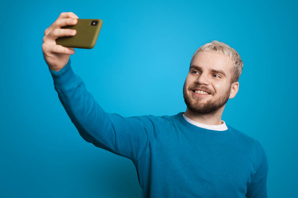 Retrato de um homem caucasiano tomando uma selfie enquanto estava de pé e olhando para a tela de seu telefone isolado sobre fundo azul. - Foto, Imagem