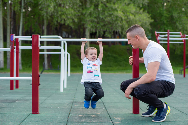 Caucasian man trains a boy on the uneven bars on the playground. Dad and son go in for outdoor sports. - Foto, Bild