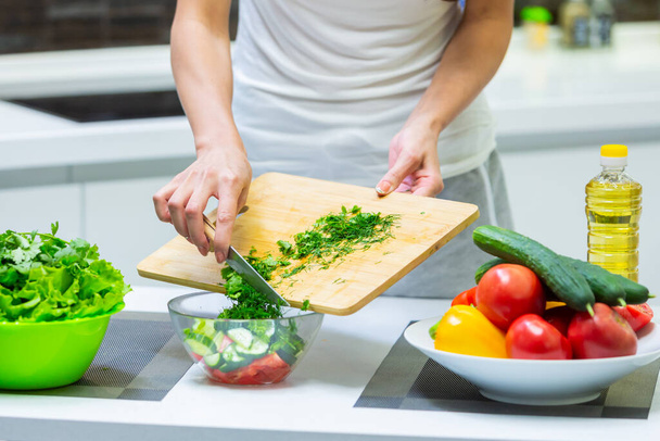 Close up of a girl hands cutting greenery with a knife on a cutting board for a vegan vitamin vegetable salad and putting them in a glass bowl while cooking breakfast in the kitchen - Φωτογραφία, εικόνα