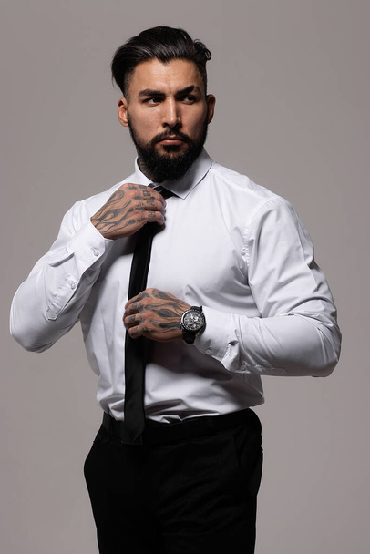 Confident Hispanic bearded man with tattooed hands in white shirt and black trousers adjusting tie and looking away while standing against gray background - Photo, Image