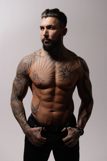 Modelo masculino hispano sin camisa con torso musculoso tatuado de pie con las manos en los bolsillos y mirando hacia otro lado sobre fondo gris - Foto, imagen
