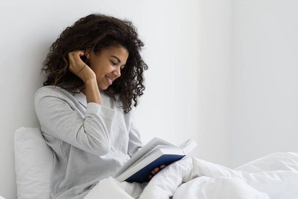 Feliz mañana en casa. Vista lateral de la joven mujer de raza mixta sonriente leyendo libro, descansando en fin de semana, sentado en la cama en pijama, pasar el día en el acogedor dormitorio blanco - Foto, imagen