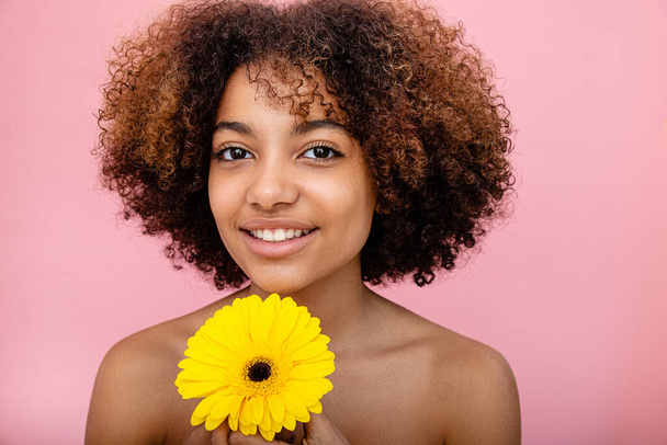 Een natuurlijk portret van een jonge donkere vrouw met krullend haar met een gele bloem in haar handen kijkend in de camera en glimlachend - Foto, afbeelding