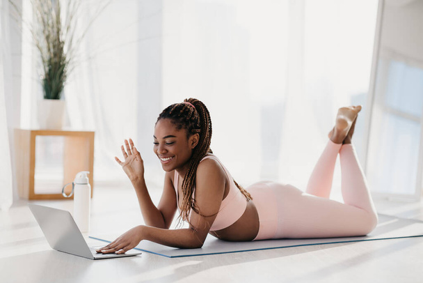 Jeune femme noire en vêtements de sport couché sur un tapis de yoga avec ordinateur portable, saluant son entraîneur personnel avant l'entraînement à la maison - Photo, image