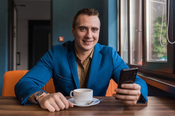 Egy fiatal, mosolygós, jóképű, európai megjelenésű üzletember, okostelefont vagy mobiltelefont használ egy kávézóban ülve egy kávézóasztalnál a kávészünetben.. - Fotó, kép