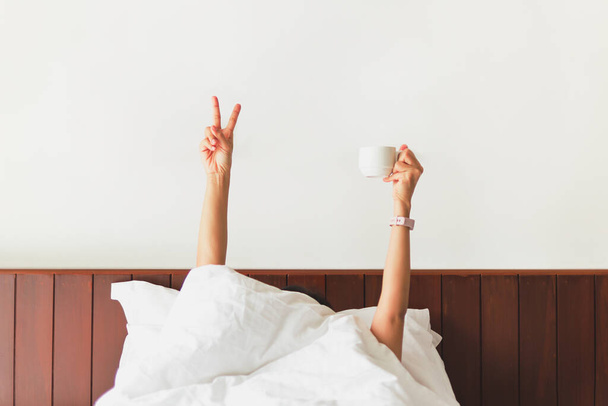 Main féminine montrant le signe drôle et tenant tasse de café derrière la couverture dans le lit. - Photo, image