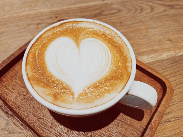 Przerwa na kawę i romantyczny nastrój. Kielich karmelowego cappuccino z piankową sztuką w kształcie serca z mleka bez laktozy, podawany na drewnianym stole w stołówce - Zdjęcie, obraz