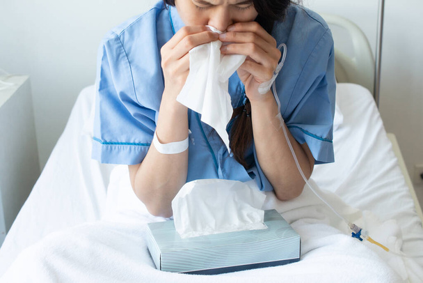 Portrait of young woman wearing patient cloths sneezing in tissue at hospital. Conceptual of sickness woman feeling when admitted in the hospital. - Photo, Image