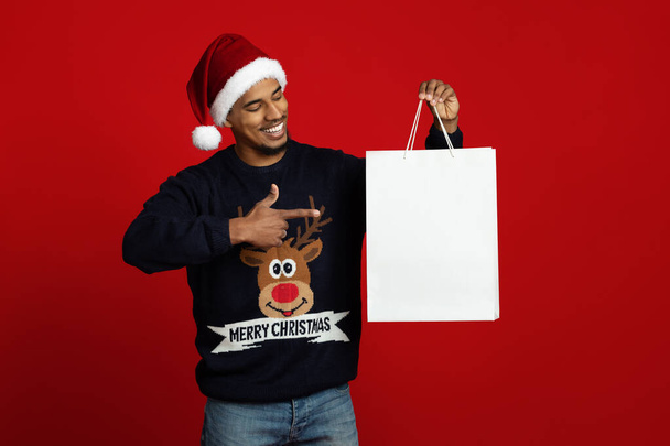 Smiling guy in Santa hat pointing at white shopping bag - Foto, Imagen