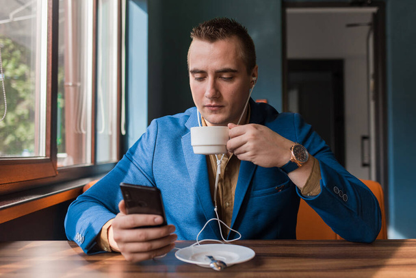 A young handsome guy businessman in blue jacket, of European appearance portrait, uses a smartphone or mobile phone sitting in a cafe at a table on a coffee break. - Φωτογραφία, εικόνα