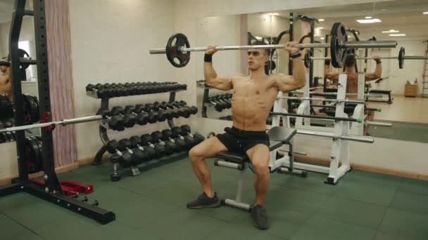 Hombre musculoso joven entrenando en el gimnasio y haciendo una barra de presión sentado en un banco para desarrollar los músculos de la cintura del hombro - Imágenes, Vídeo