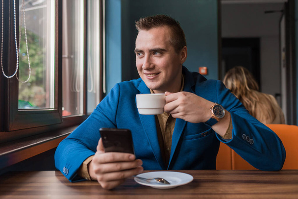 A young smiling positive handsome guy businessman of European appearance portrait, uses a smartphone or mobile phone sitting in a cafe at a table on a coffee break. - 写真・画像