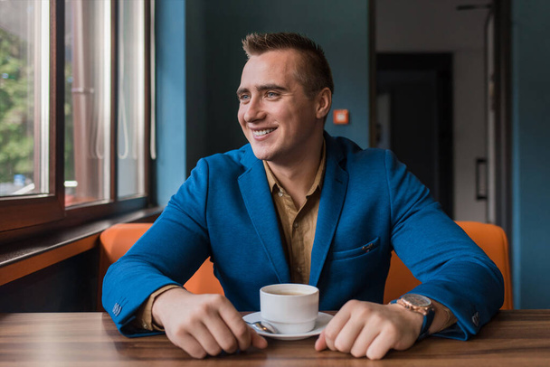 Ένας κομψός, θετικός επιχειρηματίας καυκάσιας εμφάνισης, ένας άνδρας με σακάκι και πουκάμισο κάθεται σε ένα τραπέζι σε ένα καφέ σε ένα διάλειμμα για καφέ και κοιτάζει μακριά ή έξω από το παράθυρο. - Φωτογραφία, εικόνα