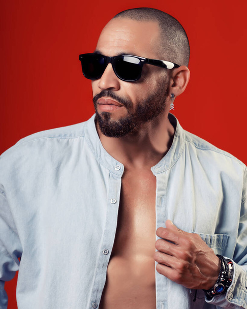 Selbstbewusster, sexy Mann in Hemd und Sonnenbrille posiert auf rotem Hintergrund. Werbe-Hintergrund Freizeitkleidung. - Foto, Bild