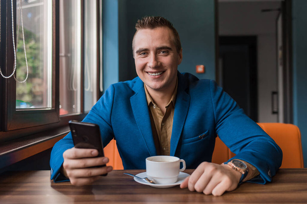 Egy fiatal, mosolygós, jóképű, európai megjelenésű üzletember, okostelefont vagy mobiltelefont használ egy kávézóban ülve egy kávézóasztalnál a kávészünetben.. - Fotó, kép