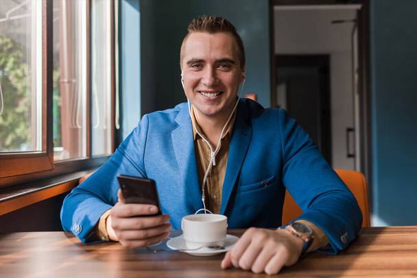 Ένας κομψός τύπος χαρούμενος χαμογελαστός επιχειρηματίας ευρωπαϊκής εμφάνισης με σακάκι και πουκάμισο κάθεται σε ένα τραπέζι σε ένα καφέ σε ένα διάλειμμα, κρατά ένα smartphone ή κινητό τηλέφωνο στο χέρι του. - Φωτογραφία, εικόνα
