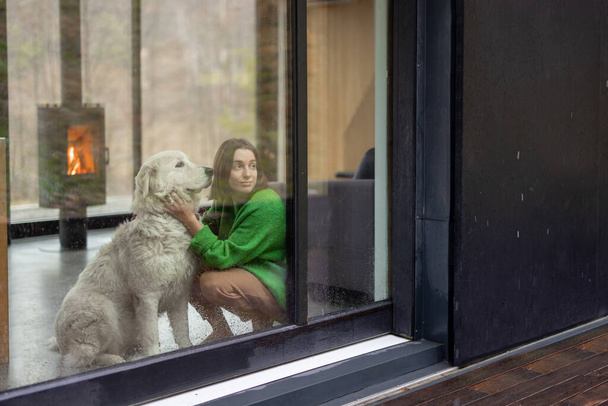 Νεαρή γυναίκα με το σκύλο της στο σύγχρονο σπίτι στη φύση - Φωτογραφία, εικόνα